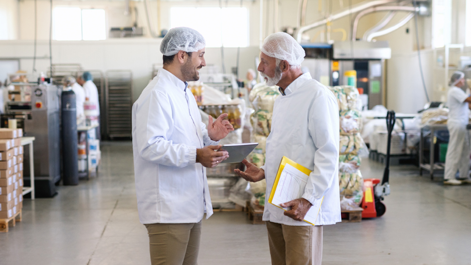 Two male food safety technicians standing and reviewing a tablet, inside a food manufacturing facility Packages of peanut-free product on a conveyor belt in a food processing facility